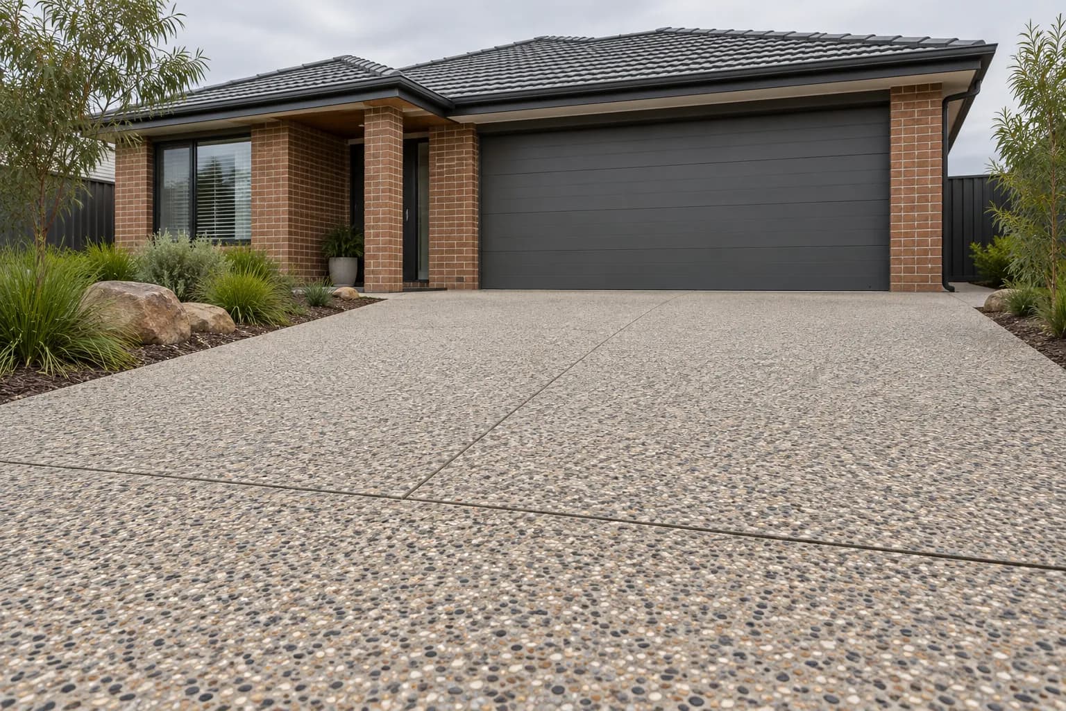 Architectural driveway detail with exposed aggregate, restrained planted border and brick home, morning side light, no workers or claim of completed work